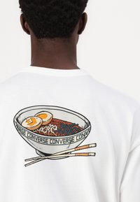 Hvit t-skjorte med et fargerikt motiv av en bolle nudler med egg og spisepinner på baksiden, med "Converse"-teksten som omkranser bollen.