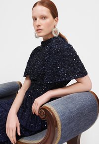 Vestido de lentejuelas azul marino con un escote alto y mangas onduladas. La modelo lleva pendientes grandes y llamativos, sentada en una silla tapizada.