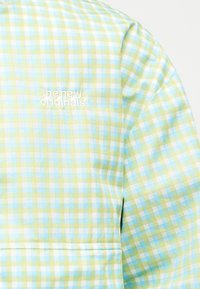 Hellgrünes, blaues und weiß kariertes Stoffmuster mit weißer Stickerei, die auf der Brust eines Kleidungsstücks den Text "the new originals" zeigt.