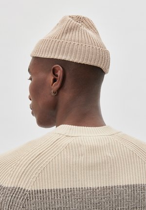 Uomo che indossa un berretto a maglia beige e un maglione beige e marrone a coste, mostrato di lato e di schiena contro uno sfondo neutro.