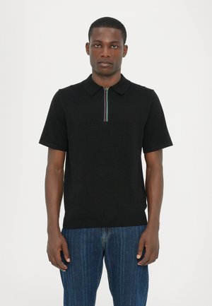 Sort kortærmet polo med lynlås og en flerfarvet stribe, lavet af glat stof, sat sammen med blå denimjeans.