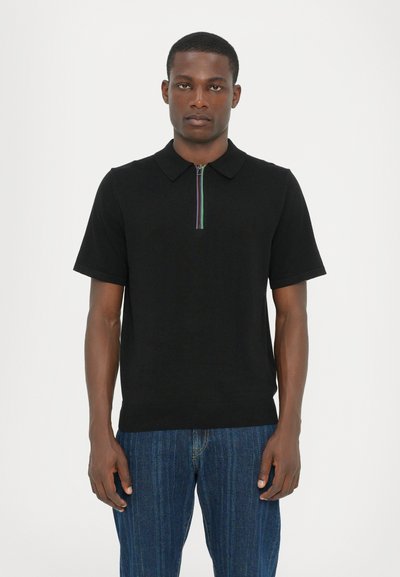 Sort kortærmet polo med lynlås og en flerfarvet stribe, lavet af glat stof, sat sammen med blå denimjeans.