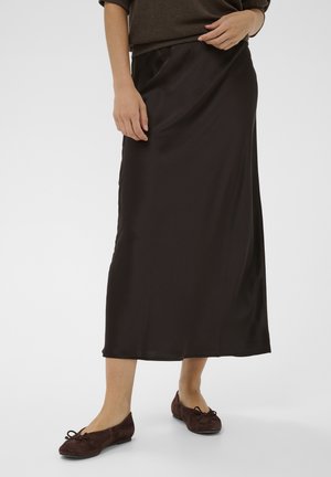 Femme portant une longue jupe marron foncé, des ballerines assorties et un haut marron à manches longues, debout devant un fond uni.