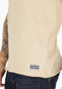 Beige cotton T-shirt with a curved hem, featuring a black and white label at the bottom left, paired with dark blue denim jeans.