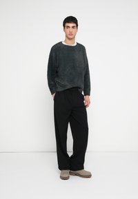 Sivý nadýchaný sveter s okrúhlym výstrihom, čierne plisované nohavice a sivé slip-on topánky. Model stojí pred jednoduchým bielym pozadím.