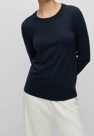 Frau trägt einen schlichten marineblauen Pullover mit langen Ärmeln und weiße Hose, steht vor einem hellgrauen Hintergrund, Hände hinter dem Rücken.