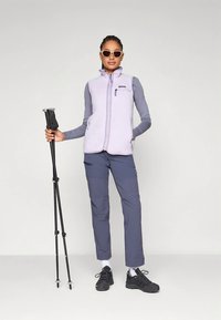 Άτομο που φοράει γυαλιά ηλίου, ανοιχτό μοβ αμάνικο γιλέκο, γκρίζο μακρυμάνικο πουκάμισο, μπλε παντελόνι, μαύρα παπούτσια πεζοπορίας, κρατώντας δύο μαύρες μπαστούνες πεζοπορίας.