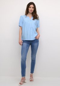 Jasnoniebieska bluzka z krótkim rękawem i dekoltem w kształcie litery V, z marszczeniami, w parze z obcisłymi niebieskimi dżinsami i jasnymi sandałami na sznurków.