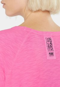 Eine blonde Person trägt ein leuchtend pinkes Hemd mit einem vertikalen Etikett auf dem oberen Rücken, auf dem steht: "SENSES THE LABEL PURE SELECTION."