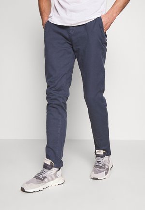 Homme portant un pantalon slim bleu marine et des baskets Adidas grises, debout les mains dans les poches devant un fond blanc.