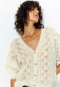 Cardigan créme texturé à coupe lâche, avec un col en V, fermeture à boutons et motifs en tricot ajouré complexes sur les manches et le corps.