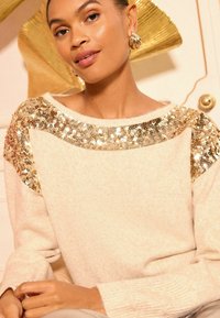 Ženska z zlatimi obročastimi uhani, oblečena v kremasto pulover s zlatimi bleščečimi detajli ob ramenih in izrezih.