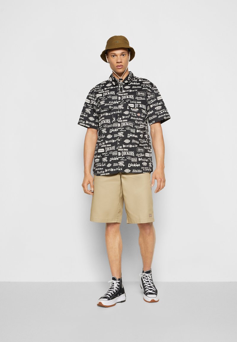 Short-sleeve black shirt with white logos, paired with tan shorts. Model wears a brown bucket hat and black high-top sneakers.