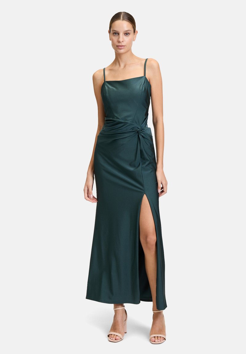 Satin, dunkelgrünes Kleid mit dünnen Trägern, tailliertem Oberteil, verdrehtem Details an der Seite und einem hohen Schlitz bis zum Oberschenkel. Getragen mit hautfarbenen, hohen Sandalen.