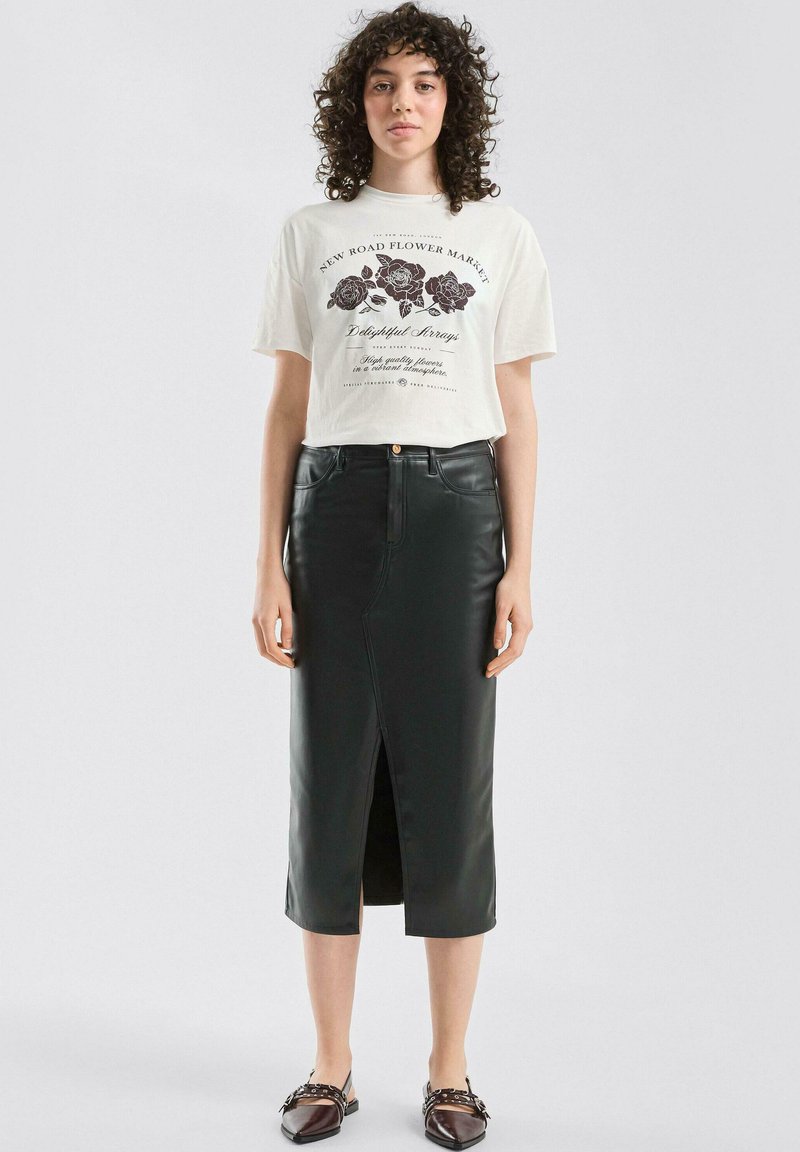 Czarna spódnica midi ze sztucznej skóry z przednim rozcięciem, zestawiona z kremowym t-shirtem z nadrukiem kwiatowym i tekstem. Look dopełniają płaskie buty.