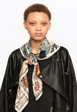 Joven con cabello corto y rizado que lleva una bufanda de patrón multicolor y una chaqueta de cuero negra sobre una camiseta oscura, con un fondo liso.