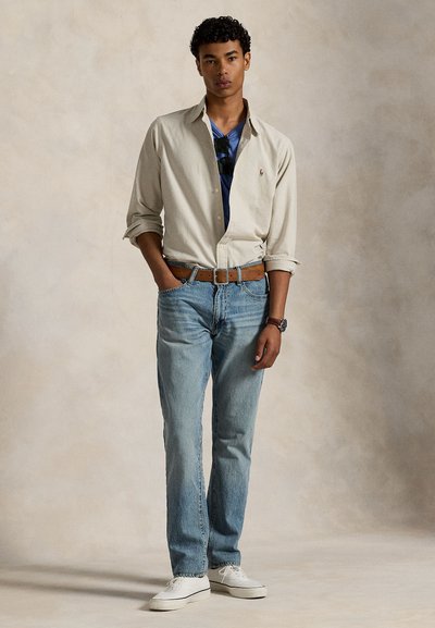 Camisa bege claro de botões por cima de uma t-shirt azul, combinada com jeans azul claro e ténis brancos. Cinto castanho e relógio completam o conjunto.