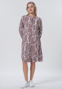 Bruine jurk met een patroon en een witte abstracte print, voorzien van een knoopsluiting aan de voorkant en een gelaagde rok. Draag het met witte sneakers.