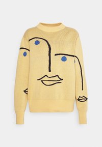 Gul stickad tröja med abstrakt svart linjekonst av ansikten och blå prickar på ögonen, med hög rund halslinning och långa ärmar.