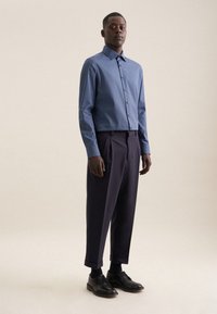 Camicia a maniche lunghe con motivo blu, abbinata a pantaloni plettati scuri e scarpe di pelle nera. Design semplice e silhouette aderente.