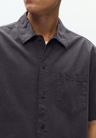 Mees, kes kannab tumedat süsi tooni nööbitavat särki rinnatasku, krae ja lühikeste varrukatega, taustal hele ühevärviline taust.