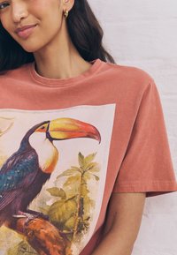Woman wearing a muted pink t-shirt with a colorful toucan perched on a branch printed on the front, standing against a light brick wall.