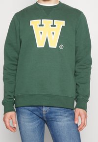 Zelená mikina s kulatým výstřihem, se na přední straně nachází velké žluté logo "W". Měkká tkanina, žebrované manžety a lem. Neformální design.