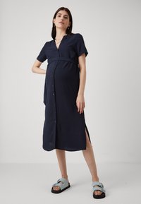 Ναυτικό μπλε φόρεμα με κουμπιά και κοντά μανίκια, πτυχωτή υ�φή, ζωσμένο στη μέση και με πλαϊνές σχισμές, σε συνδυασμό με ανοιχτό μπλε σανδάλια.