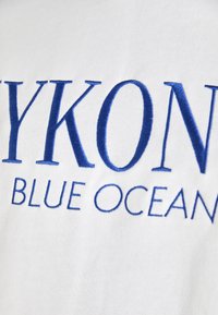 Bela tkanina z modrim vezenim besedilom, ki pravi "MYKONOS BLUE OCEAN." Preprost, neopazen dizajn s jasnimi in razločno napisanimi črkami.