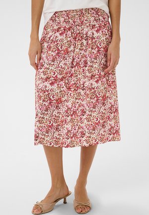 Kobieta ubrana w różową, czerwoną i brązową, kwiecistą spódnicę midi, do której dobrała beżowe sandały na niskim obcasie z odkrytymi palcami.