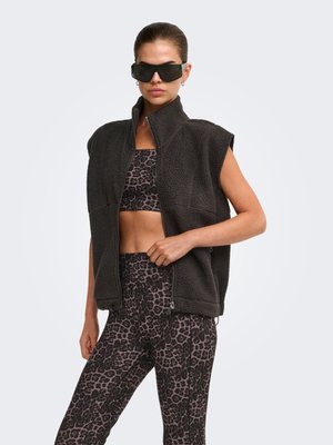 Femme portant une veste polaire noire sans manches sur un soutien-gorge de sport imprimé léopard et un legging, avec de grandes lunettes de soleil foncées.