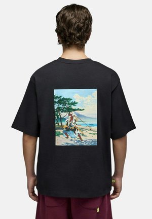 Personne aux cheveux bouclés portant un t-shirt noir avec un imprimé coloré style ukiyo-e représentant un samouraï brandissant un bâton en plein air.