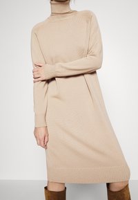 Robe beige en tricot à manches longues et col roulé portée avec des bottes marron en daim montantes jusqu'aux genoux, un bras croisé sur le corps, fond gris uni.