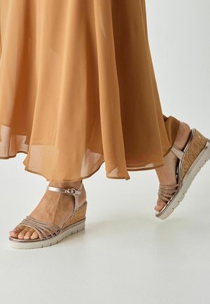 Keilsandalen mit Korkabsatz, metallischen Riemen und einer weichen beigefarbenen Sohle. Ein schmaler Knöchelriemen mit einer Schnallendetail vervollständigt das Design.