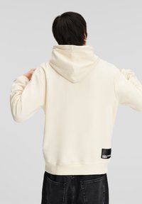 Felpa leggera color crema con cappuccio regolabile, tessuto morbido, orlo a coste e una toppa nera con logo sul lato sinistro inferiore.