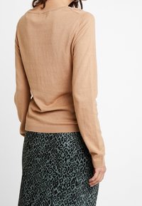 Maglione lungo a maniche con un morbido tessuto a maglia di colore marrone chiaro, abbinato a una gonna con stampa leopardata verde. Collo a giro semplice e orlo a costine.