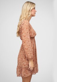 Robe fluide à fleurs marron avec de longues manches bouffantes, un corsage smocké et un ourlet en volants. Présente de petits motifs de fleurs rouges et blanches.