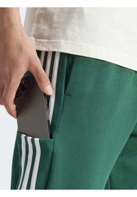 Gröna sweatpants med vita ränder på sidorna, som har en ficka som håller en smartphone. Tyget verkar texturerat och avslappnat.
