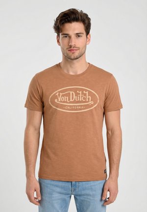 Joven con cabello castaño ondulado que lleva una camiseta marrón claro con el logo de Von Dutch California y jeans azul claro, de pie frente a un fondo liso.