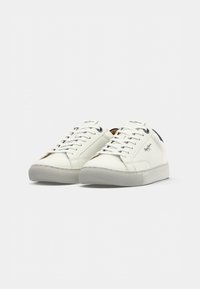 Paire de baskets basses blanches en cuir avec lacets et semelles en caoutchouc gris sur un fond gris clair uni.
