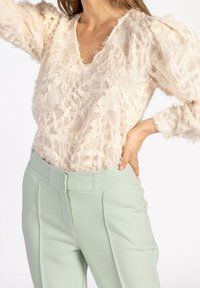 Donna che indossa una blusa a maniche lunghe di crema testurizzata con spalle a palloncino e pantaloni a vita alta verde chiaro, mano sui fianchi.