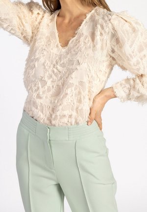 Frau in einer strukturierten, cremefarbenen Bluse mit langen Ärmeln und puffigen Schultern, die hellgrüne, hochgeschnittene Hose trägt und die Hand auf die Hüfte legt.