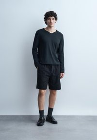 Młody mężczyzna stoi przy jasnej ścianie, ubrany w czarną koszulę z długim rękawem, czarne szorty w wzory, czarne skarpetki i czarne skórzane buty.