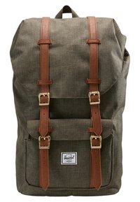 Mochila de tela verde oliva con correas de cuero marrón, hebillas doradas, bolsillo frontal y parche rectangular blanco con el logo de Herschel.