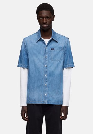 Mężczyzna-model ubrany w niebieską, krótko rękawową koszulę jeansową nałożoną na białą, długorękawową koszulę, w czarnych spodniach, stojący na białym tle.