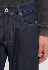 Mørkeblå denimjeans med rett snitt, synlig søm, metallknapp-lukking og et lite gult merke nær lommen.