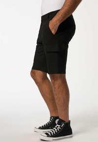 Schwarze Cargo-Shorts mit Seitentaschen und geradem Schnitt, kombiniert mit hochschuligen schwarzen Canvas-Sneakern mit weißen Sohlen und Ösen.