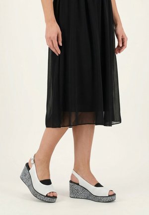 Beine mit weißen perforierten Keilsandalen mit gemusterter Plateausohle und einem schwarzen halbtransparenten Midirock.