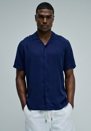 Hombre con camisa azul marino de manga corta con botones y pantalones blancos con cordón, de pie con las manos en los bolsillos contra un fondo liso.