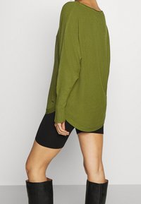 Pull en tricot vert olive, coupe ample, avec un ourlet arrondi, des manches longues et un tissu texturé. Associé à un short noir et des bottes hautes noires.